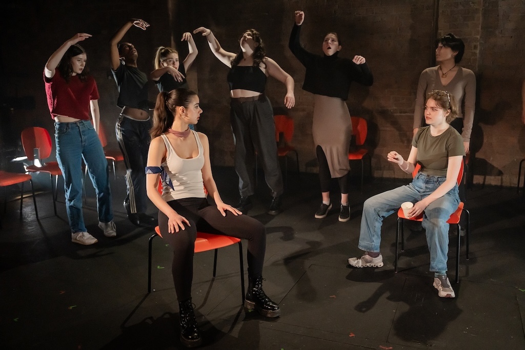 Action shot of the spring 2024 Shakespeare in Performance at RADA final presentation of Julius Caesar. Two actors sit in bright orange chairs while other actors stand behind them with arms above their heads and concerned expressions on their faces.